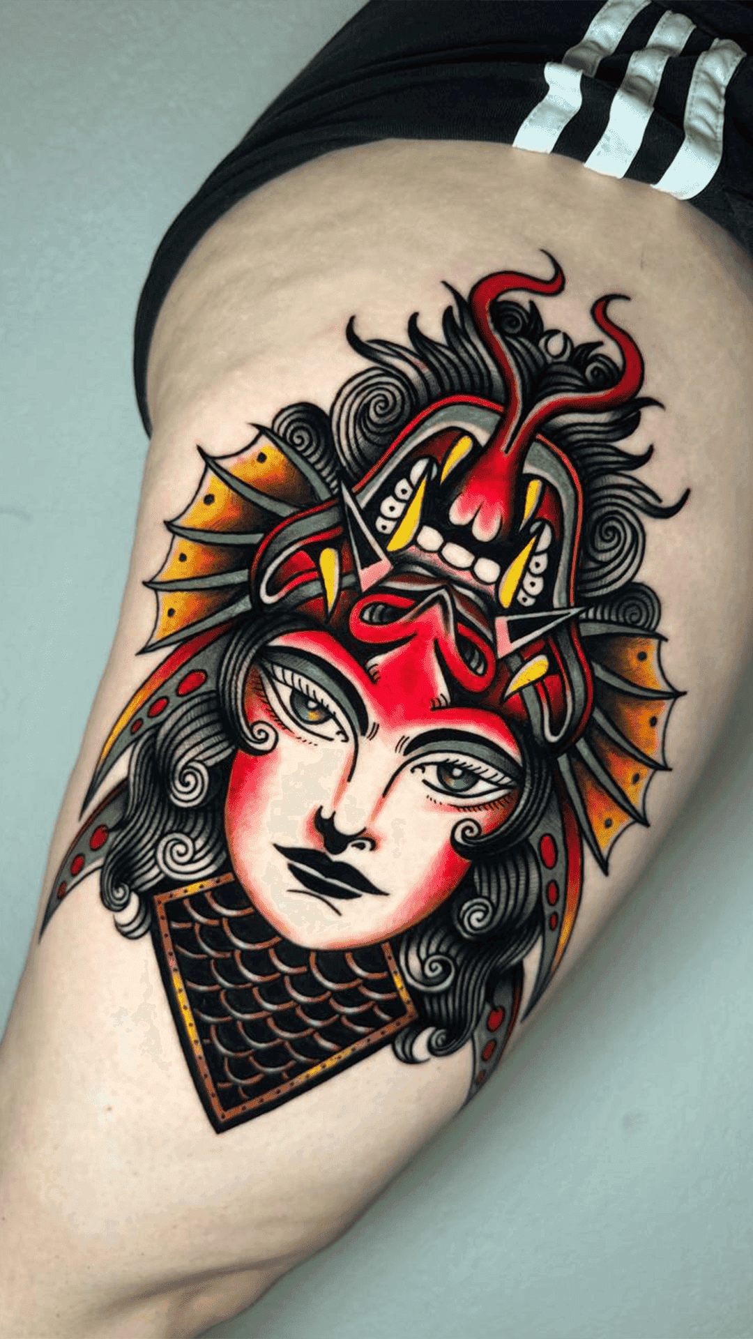 Traditional-style color tattoo of a woman's face crowned with a red dragon headdress, on the thigh