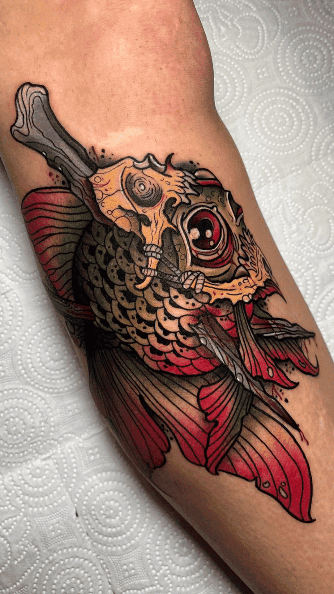 Neo-traditional color tattoo of a red-finned fish with a skull face and a bone, on a forearm